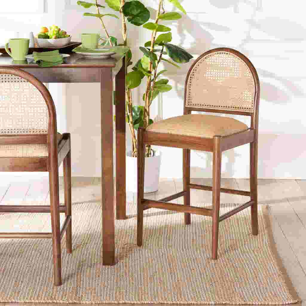 A wooden counter stool with a woven cane back and seat, placed at a dining table with green cups and a plant in the background.